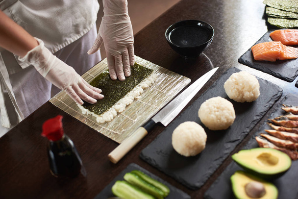 Vista de perto das mãos do chef preparando sushi rolante com ingredientes: arroz, nori, abacate, salmão, molho de soja
 - Foto, Imagem