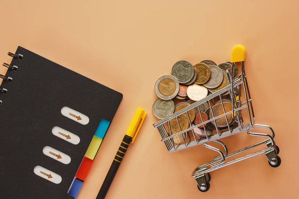 Money and coins in mini shopping cart or trolley with spiral notebook and pen on beige color background for spending plan, investment, business and finance concept - Foto, imagen