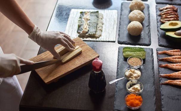 Close-up weergave van het proces ter voorbereiding van rollende sushi. Handen in wegwerphandschoenen snijden omelet op een houten bord - Foto, afbeelding