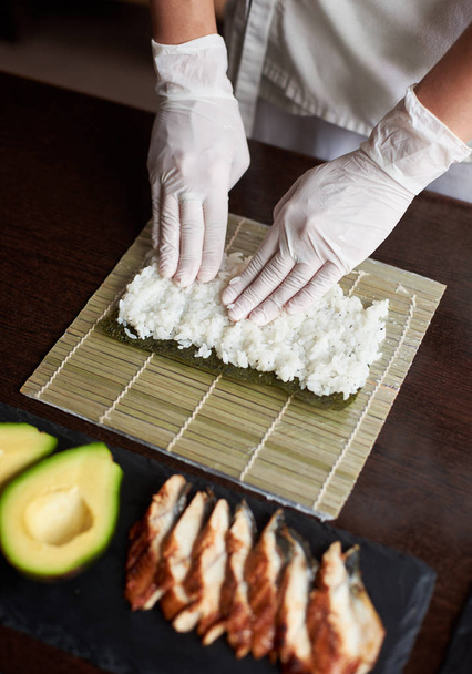 Vista de perto do processo de preparação de sushi de rolamento. Nori e arroz branco em esteira de bambu. As mãos do Chef tocam arroz. Chef começa a cozinhar sushi. Enguia e abacate
 - Foto, Imagem