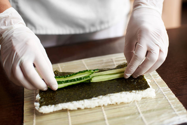 Close-up weergave van het proces ter voorbereiding van rollende sushi. Nori, witte rijst en komkommer op bamboe mat. Chef's handen in handschoenen begint koken sushi rolls - Foto, afbeelding