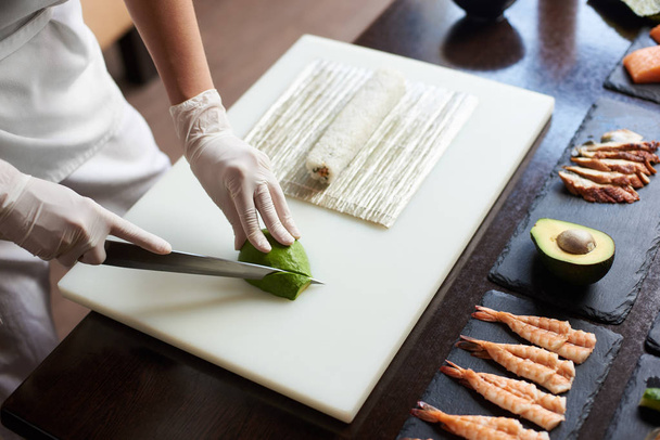 Vista de perto do processo de preparação de delicioso sushi rolante no restaurante. Mãos femininas em luvas descartáveis cortando abacate em tábua de madeira com a faca
. - Foto, Imagem