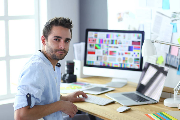 Portrait de jeune designer assis dans un studio graphique devant un ordinateur portable et un ordinateur tout en travaillant en ligne. - Photo, image