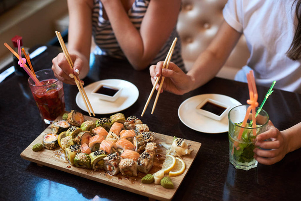 Two yang girls are eating delicious rolling sushi at restaurant served on the wooden board with chopsticks, soy sauce and cocktails - Photo, Image