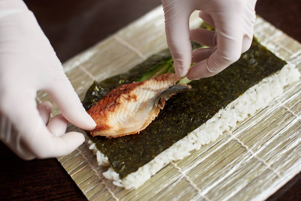 Close-up weergave van het proces ter voorbereiding van rollende sushi. Nori, witte rijst, komkommer en paling op bamboe mat. Chef's handen in handschoenen begint koken sushi rolls - Foto, afbeelding