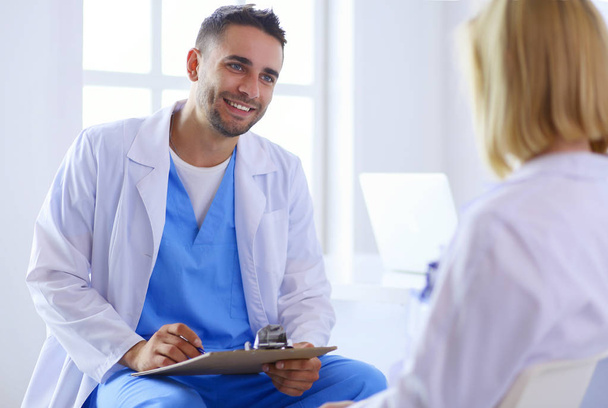 Handsome doctor is talking with young female patient and making notes while sitting in his office - Foto, imagen