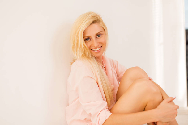 Ευτυχής όμορφη νεαρή ξανθιά κοπέλα με ένα χαμόγελο σε ένα ροζ πουκάμισο κάθεται στο πάτωμα - Φωτογραφία, εικόνα