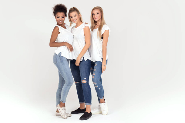 Grupo de jóvenes muchachas atractivas multiétnicas con camisas blancas, sonriendo y divirtiéndose juntas, posando en el estudio
. - Foto, Imagen