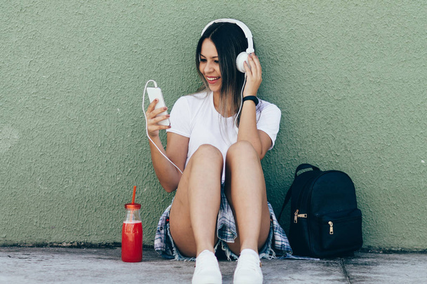 Portrait of a happy young woman listening to music via headphones on the street - Фото, изображение