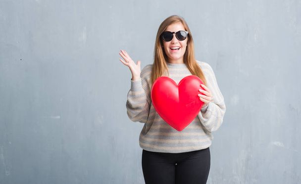Young adult woman over grey grunge wall holding red heart in love very happy and excited, winner expression celebrating victory screaming with big smile and raised hands - Photo, Image