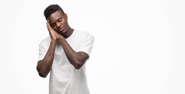 Νεαρός αφρικανική αμερικανική άνθρωπος που φορώντας άσπρο t-shirt στον ύπνο κουρασμένος όνειρα και ποζάρουν με τα χέρια μαζί ενώ χαμογελώντας με κλειστά τα μάτια. - Φωτογραφία, εικόνα