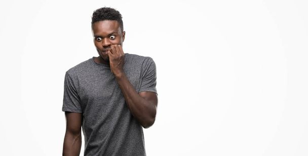 junger afrikanisch-amerikanischer Mann in grauem T-Shirt, der gestresst und nervös aussieht, die Hände auf den Mund beißenden Nägeln. Angstproblem. - Foto, Bild