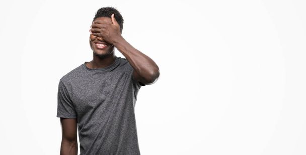 Молодой африканский американец в серой футболке улыбается и смеется, закрывая глаза рукой. Слепая концепция
. - Фото, изображение