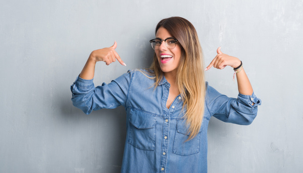 Jovem mulher adulta sobre grunge parede cinza usando óculos olhando confiante com sorriso no rosto, apontando-se com os dedos orgulhosos e felizes
. - Foto, Imagem