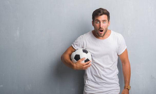 Przystojny młody mężczyzna ponad szary tło ściany trzymając piłki nożnej Piłka nożna przestraszony w szoku ze ścianą niespodzianka, boi się i podekscytowany z wyrazem strachu - Zdjęcie, obraz