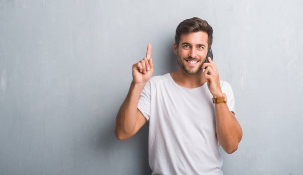 Handsome young man over grey grunge wall speaking on the phone surprised with an idea or question pointing finger with happy face, number one - Фото, изображение
