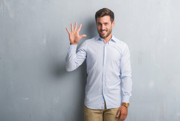 schöner junger Geschäftsmann über grauer Grunge-Wand mit elegantem Hemd, das die Finger Nummer fünf zeigt und nach oben zeigt, während er zuversichtlich und glücklich lächelt. - Foto, Bild