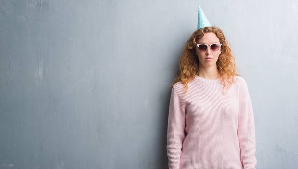 Jeune femme rousse sur le mur gris grunge portant un chapeau d'anniversaire avec une expression confiante sur la pensée intelligente du visage grave
 - Photo, image