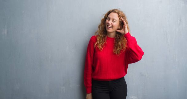 Mladá zrzka žena nad šedá grunge zdi nosí červený svetr s úsměvem s rukou přes ucho naslouchá slyšení zvěsti nebo drby. Hluchota koncept. - Fotografie, Obrázek