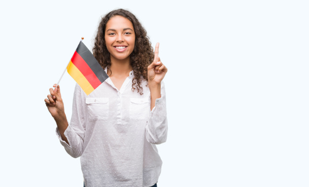 Gazdaság, Németország zászlaja meglepett mutatóujjal egy ötletet vagy kérdést boldog arc, számú fiatal spanyol nő - Fotó, kép