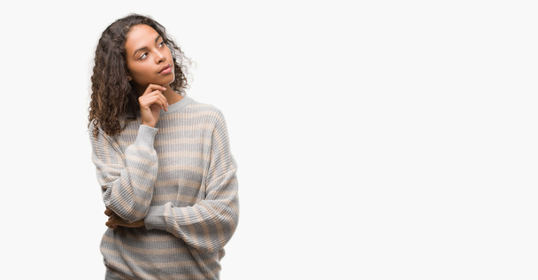 Beautiful young hispanic woman wearing stripes sweater with hand on chin thinking about question, pensive expression. Smiling with thoughtful face. Doubt concept. - Photo, Image