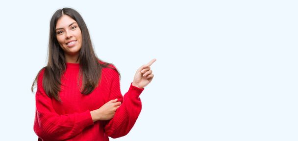 Young beautiful hispanic wearing red sweater with a big smile on face, pointing with hand and finger to the side looking at the camera. - Photo, Image