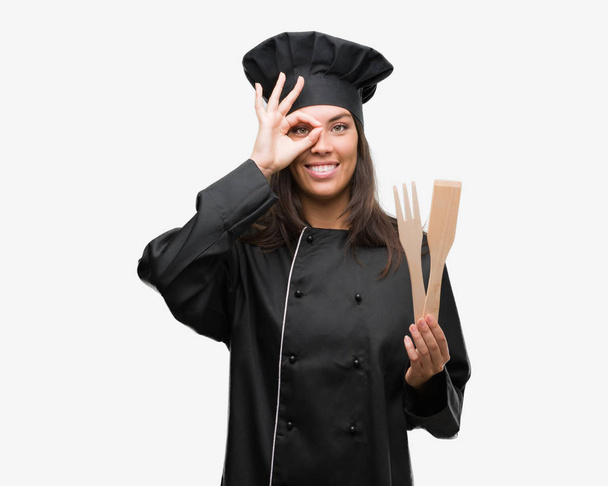Молода іспаномовна кулінарна жінка в уніформі шеф-кухаря з щасливим обличчям, посміхаючись, роблячи знак ОК з рукою на око дивиться крізь пальці
 - Фото, зображення