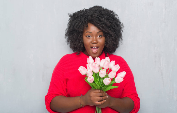 Joven mujer afroamericana sobre pared grunge gris sosteniendo ramo de flores rosadas asustada en shock con una cara sorpresa, asustada y excitada con expresión de miedo
 - Foto, imagen