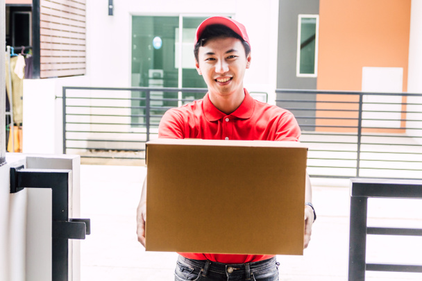Happy dodávky muž v červené uniformě držení lepenkové krabice stojí v blízkosti domu customer.kurýrní služby koncept - Fotografie, Obrázek