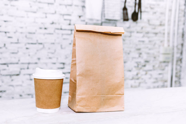 Tasse Kaffee und leere Papiertüte mit Kopierplatz für Ihre Marke auf dem Tisch vor weißem Backsteinhintergrund - Foto, Bild