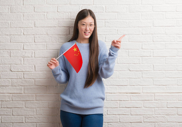 Молодая китаянка за кирпичной стеной с флагом Китая очень довольна, указывая рукой и пальцем в сторону
 - Фото, изображение