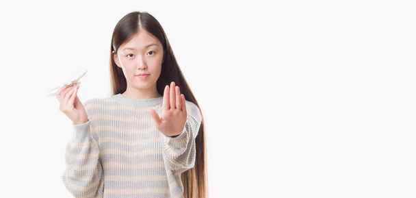 Jovem chinesa sobre fundo isolado segurando brinquedo de avião com a mão aberta fazendo sinal de parada com expressão séria e confiante, gesto de defesa
 - Foto, Imagem