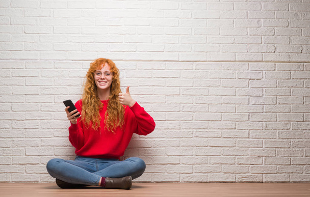 Young redhead woman sitting over brick wall talking on the phone happy with big smile doing ok sign, thumb up with fingers, excellent sign - Фото, изображение