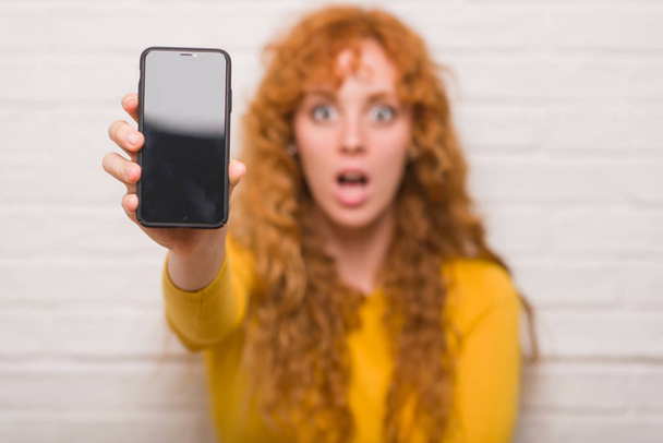 junge rothaarige Frau sitzt über Ziegelmauer und zeigt schockiertes Smartphone mit überraschendem Gesicht, ängstlich und aufgeregt mit ängstlichem Ausdruck - Foto, Bild