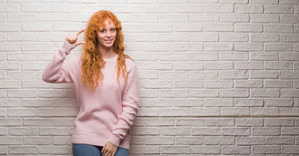 Young redhead woman standing over brick wall smiling and confident gesturing with hand doing size sign with fingers while looking and the camera. Measure concept. - Photo, Image