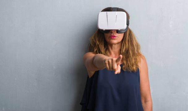 hispanische Frau mittleren Alters, die mit einer Virtual-Reality-Brille über einer grauen Grunge-Wand steht und mit dem Finger auf die Kamera und dich zeigt, Handzeichen, positive und selbstbewusste Geste von vorne - Foto, Bild