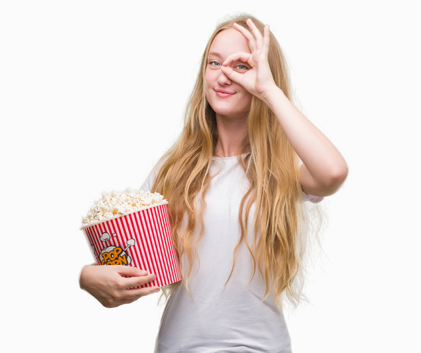 Loira adolescente mulher comer milho pop com rosto feliz sorrindo fazendo ok sinal com a mão no olho olhando através dos dedos
 - Foto, Imagem