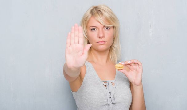 Взрослая белая женщина за гранж серой стеной ест макарон с открытой рукой делает знак стоп с серьезным и уверенным выражением лица, защитный жест
 - Фото, изображение