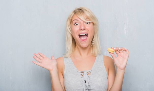 Mujer caucásica adulta sobre pared gris grunge comiendo macaron muy feliz y emocionada, expresión ganadora celebrando la victoria gritando con una gran sonrisa y levantando las manos
 - Foto, Imagen