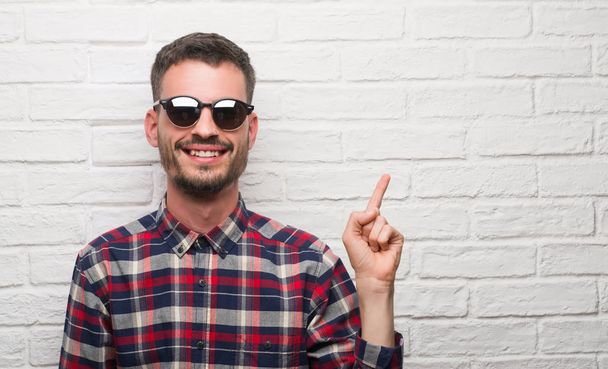 Mladý dospělý muž nosí sluneční brýle stojící nad bílé zdi velmi spokojený, s rukou a prstu přejdete na stranu - Fotografie, Obrázek