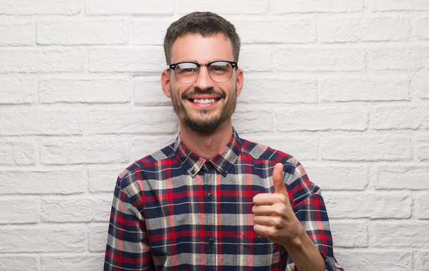 Young adult man standing over white brick wall happy with big smile doing ok sign, thumb up with fingers, excellent sign - Photo, Image