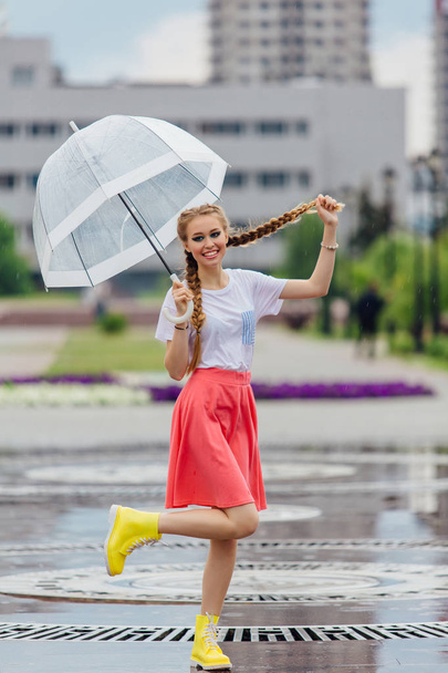 Молода красива дівчина з двома косами в жовтих чоботях і з прозорою парасолькою стоїть біля фонтану. Дощовий день у місті
. - Фото, зображення