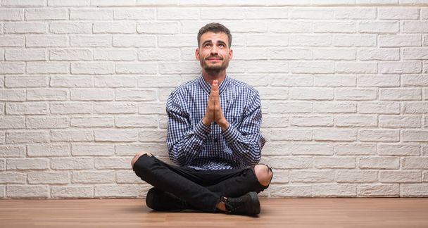 Hombre adulto joven sentado sobre la pared de ladrillo blanco mendigando y rezando con las manos junto con la expresión de esperanza en la cara muy emocional y preocupado. Pedir perdón. Concepto de religión
. - Foto, Imagen