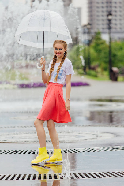Young pretty girl with two braids in yellow boots and with transparent umbrella stands near fountain. Rainy day in city. - Fotografie, Obrázek