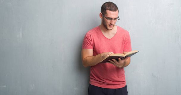 Jovem caucasiano sobre parede grunge cinza lendo um livro com uma expressão confiante no rosto inteligente pensando sério
 - Foto, Imagem
