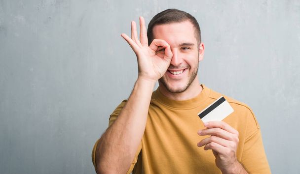 Młody człowiek kaukaski ponad szary tło ściany trzymając kartę kredytową z szczęśliwy twarz uśmiechający się robi ok znak ręką na oko patrząc przez palce - Zdjęcie, obraz