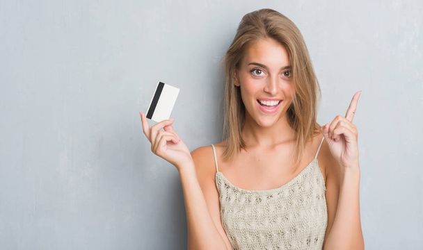 Beautiful young woman over grunge grey wall holding credit card surprised with an idea or question pointing finger with happy face, number one - Φωτογραφία, εικόνα