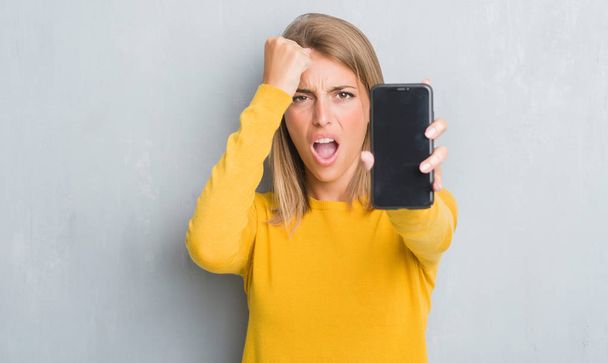 Красивая молодая женщина за гранж серой стеной показывает экран смартфона раздраженный и разочарованный кричать от гнева, сумасшедший и кричать с поднятой рукой, гнев концепции
 - Фото, изображение