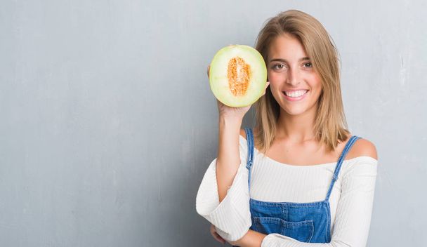 Krásná mladá žena grunge šedá zeď drží čerstvé meloun meloun s šťastný obličej stál a usmíval se s důvěřivým úsměvem ukázal zuby - Fotografie, Obrázek