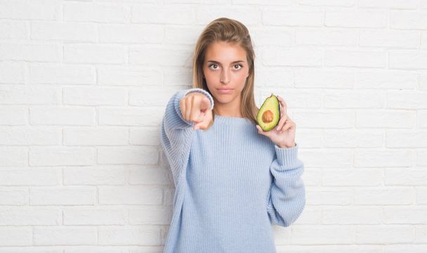 Bella giovane donna sul muro di mattoni bianchi mangiare avocado fresco puntando con il dito alla fotocamera e a voi, segno della mano, gesto positivo e fiducioso dal davanti
 - Foto, immagini